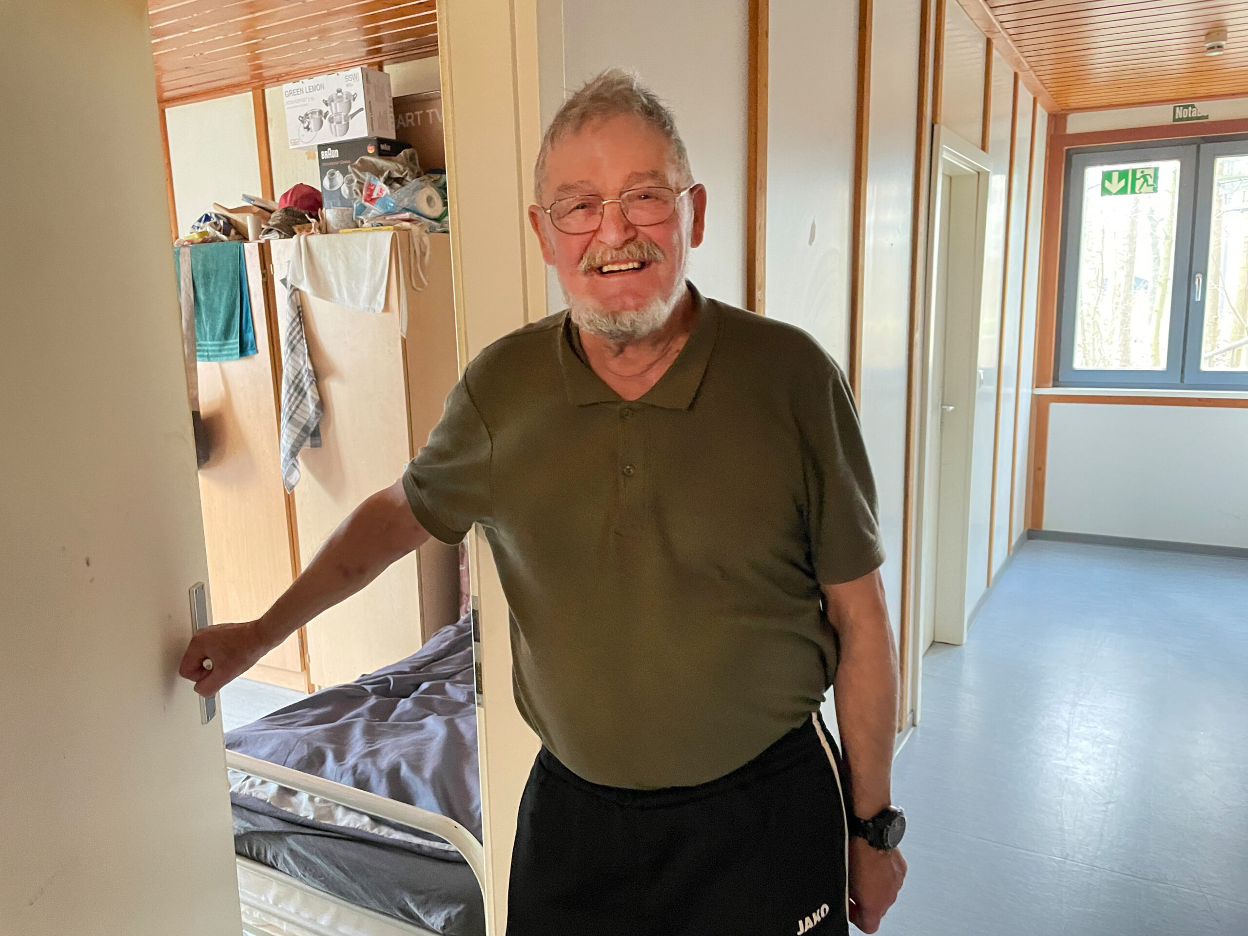 Peter, Jogginghose, olivfarbenes Polo-Shirt, steht im Flur einer Wohnunterkunft von „Fördern & Wohnen“.