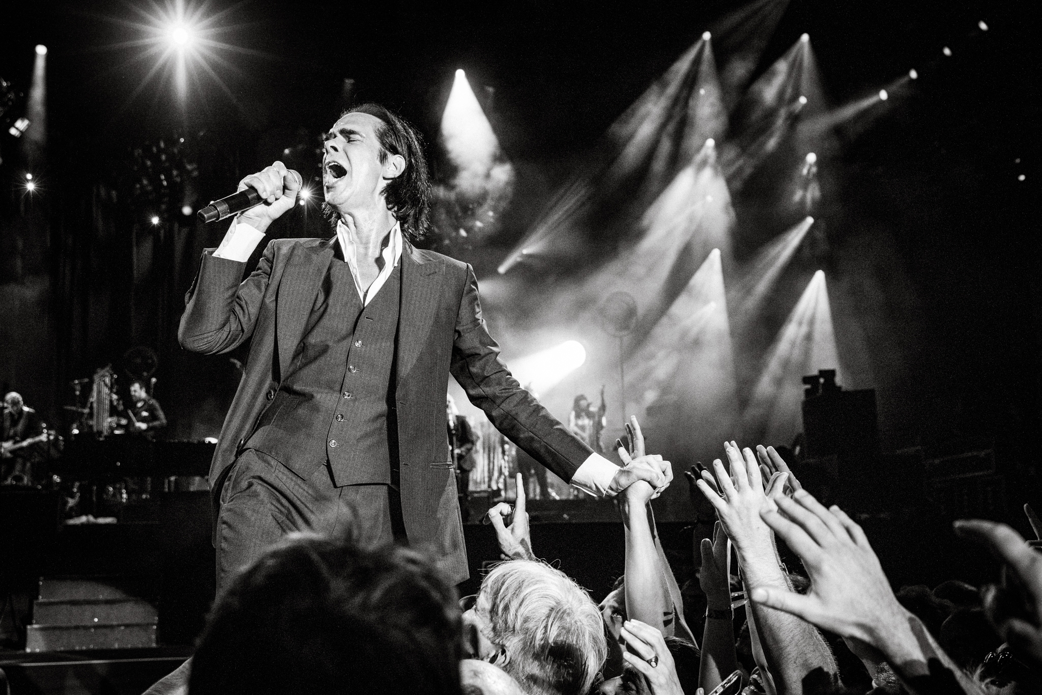 Schwarzweißfoto, Nick Cave singt ins Mikrofon, mit der anderen Hand hält er die Hand eines Fans