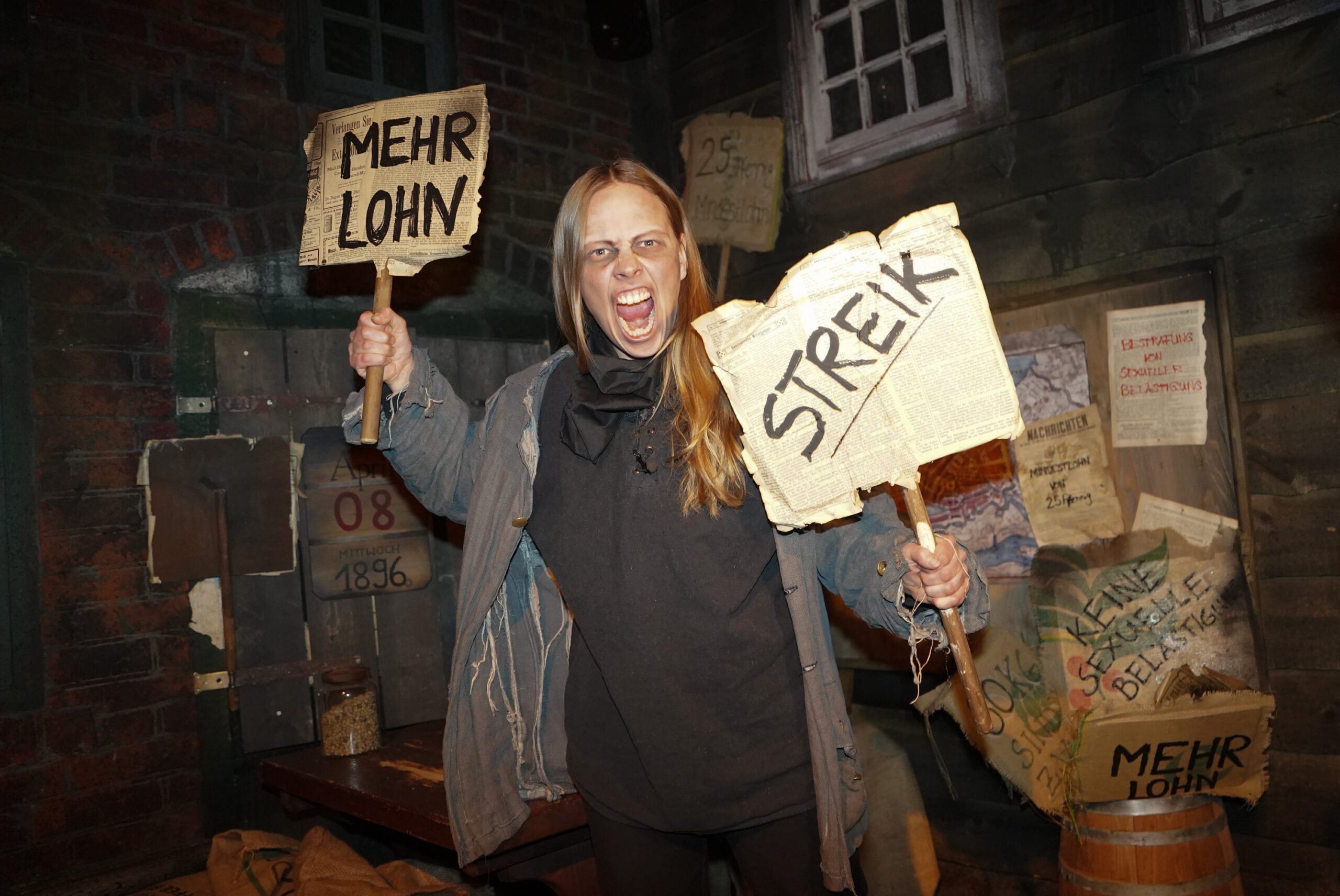 Show im Hamburg Dungeon: Eine Frau als streikende Kaffeeverleserin vor 128 Jahren; sie hält zwei Plakate hoch.