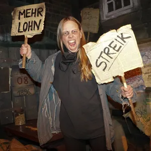 Show im Hamburg Dungeon: Eine Frau als streikende Kaffeeverleserin vor 128 Jahren; sie hält zwei Plakate hoch.