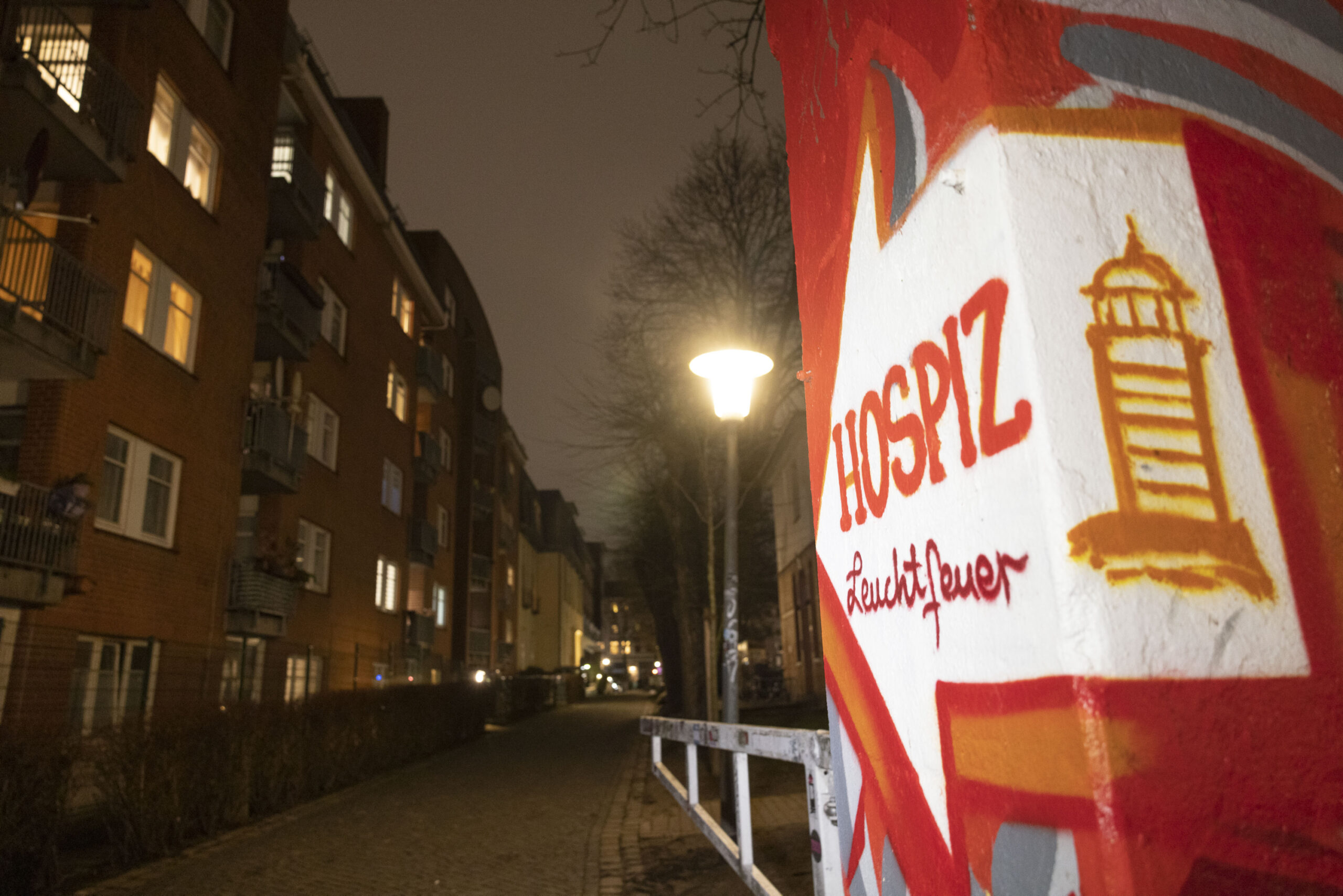 Eine rot angemalte Mauer, darauf ein weißer Pfeil, auf dem steht „Hospiz Leuchtfeuer“, daneben ein Leuchtturm