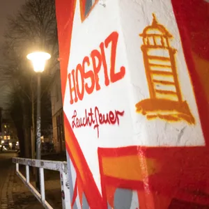 Eine rot angemalte Mauer, darauf ein weißer Pfeil, auf dem steht „Hospiz Leuchtfeuer“, daneben ein Leuchtturm