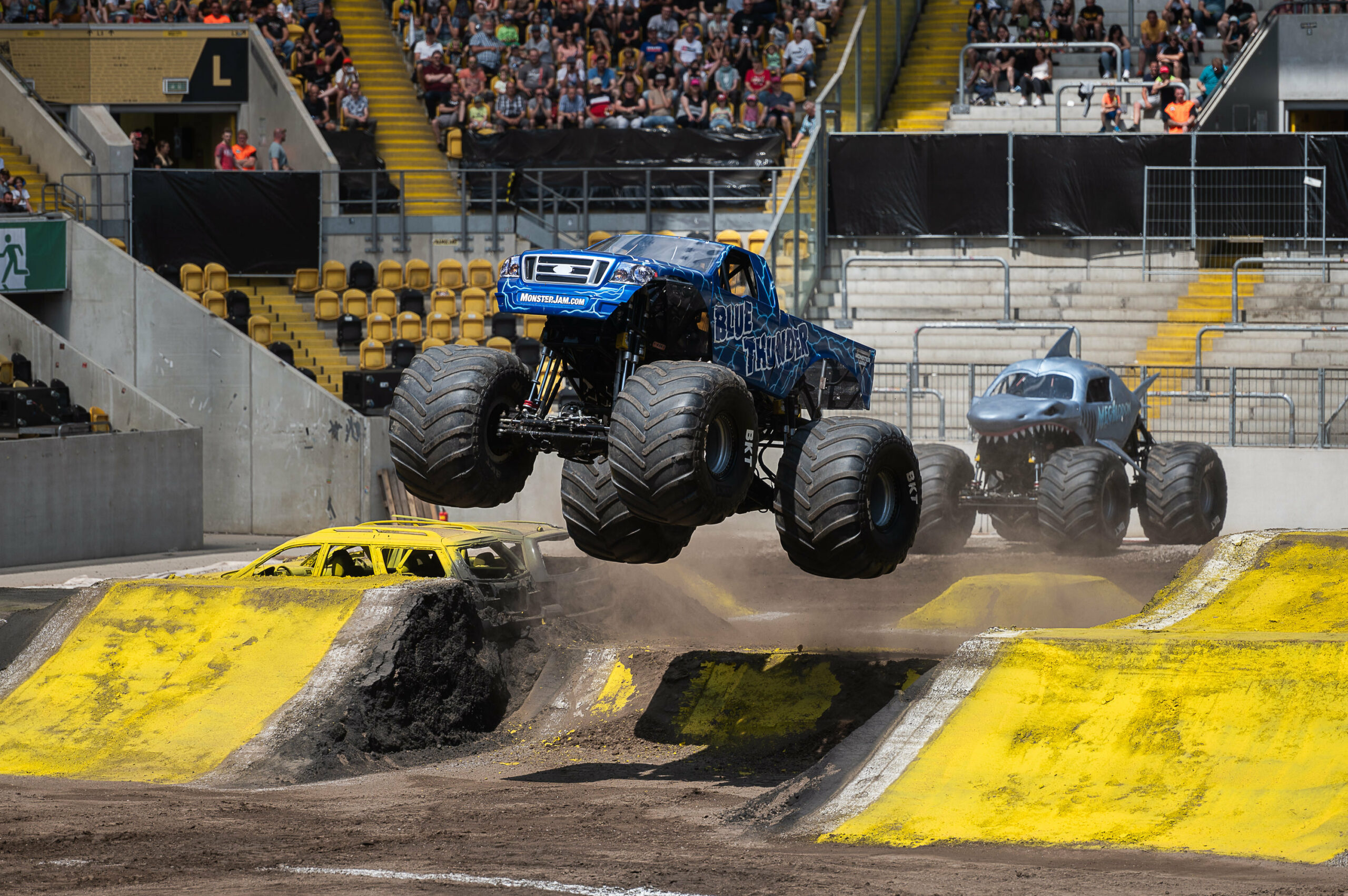 Der Monster Truck „Blue Thunder“ springt über eine Rampe bei der Show „Monster Jam“, dahinter ein Monster Truck, der aussieht wie ein Hai