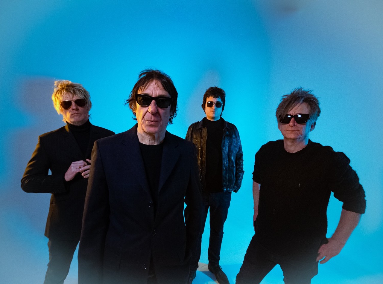 Vier Bandmitglieder vor blauem Hintergrund, alle mit Sonnenbrillen