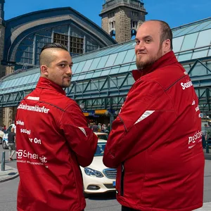 Die Sozialraumläufer Kevin (l.) und Karsten stehen vor dem Hauptbahnhof, sie tragen rote Jacken.