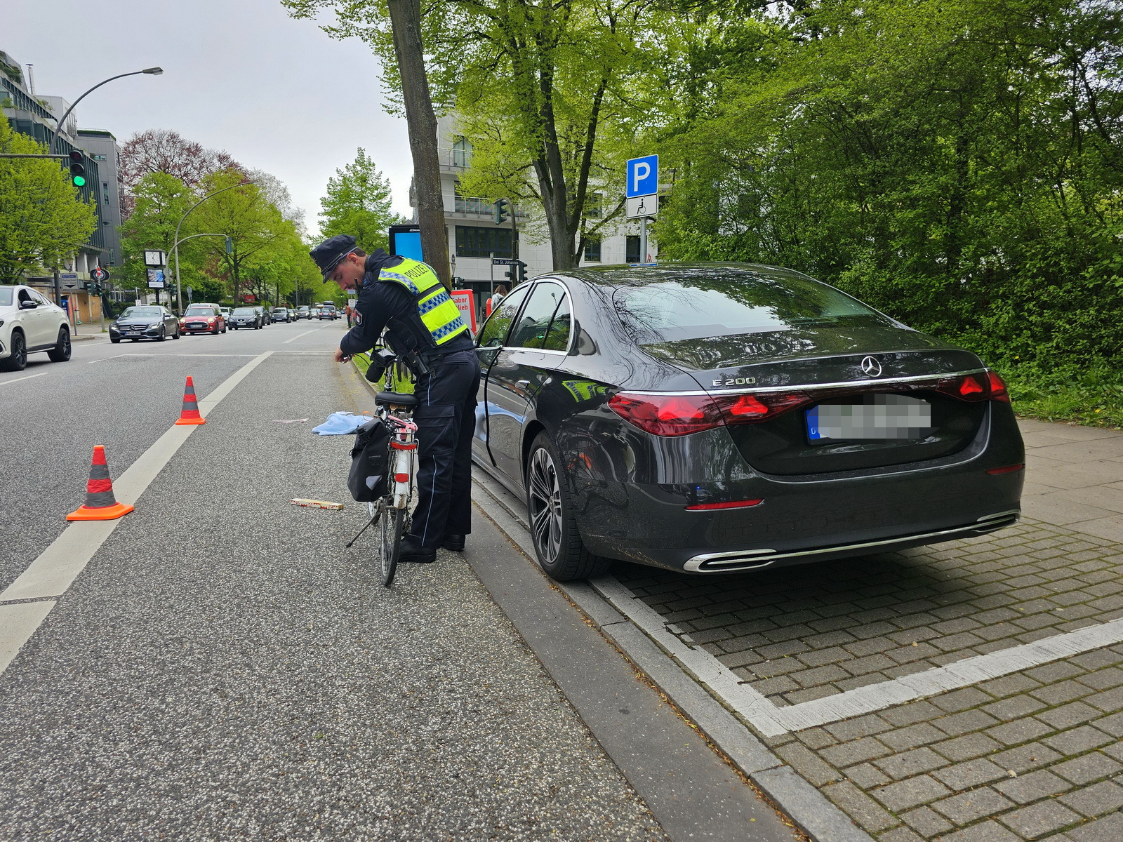 Ein Polizist untersucht das Fahrrad der verunglückten Frau, daneben auf dem Parkstreifen steht eine schwarze Mercedes-Limousine.