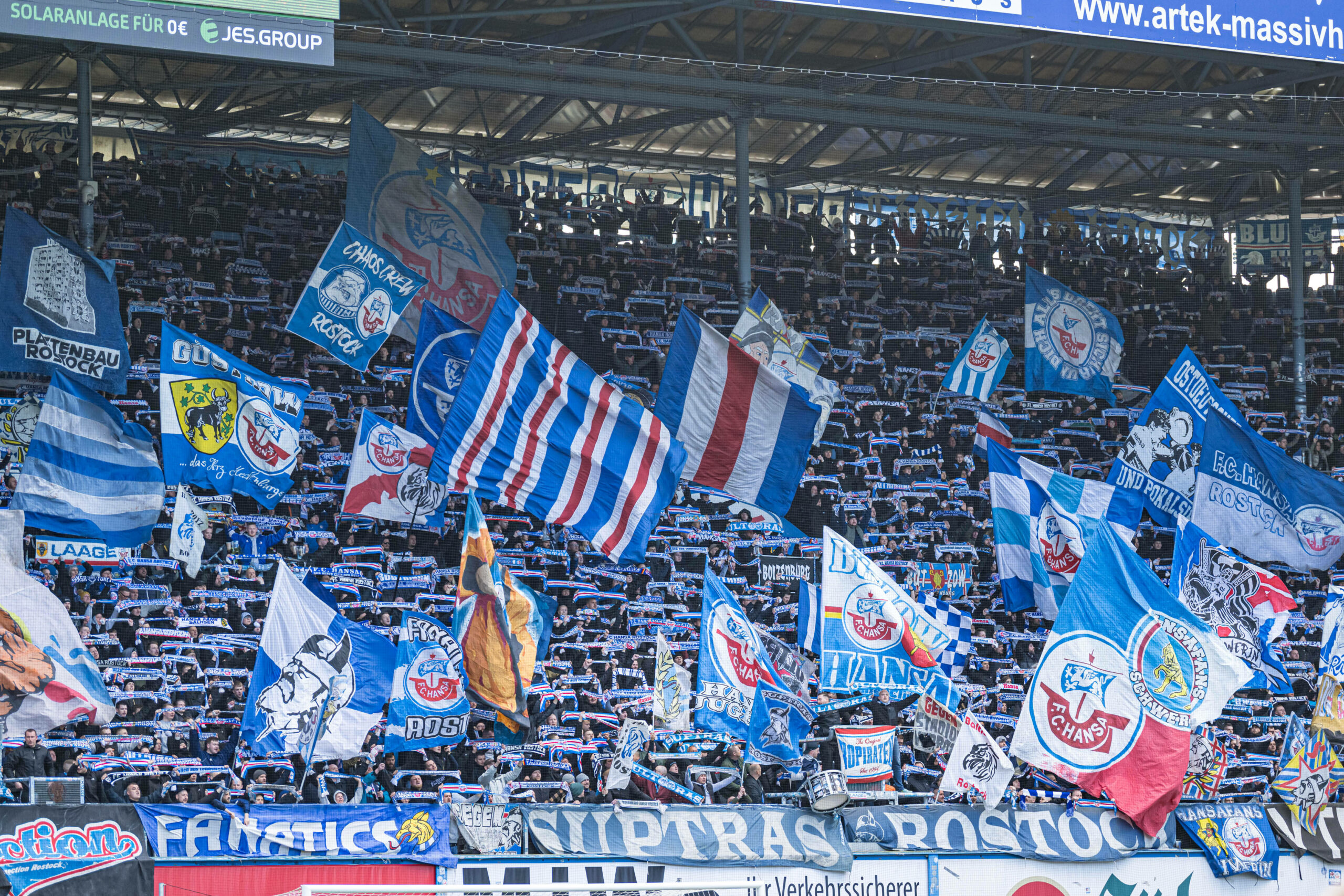 Die Fans von Hansa Rostock schwenken die Fahnen und halten ihre Schals in die Luft bei einem Spiel.