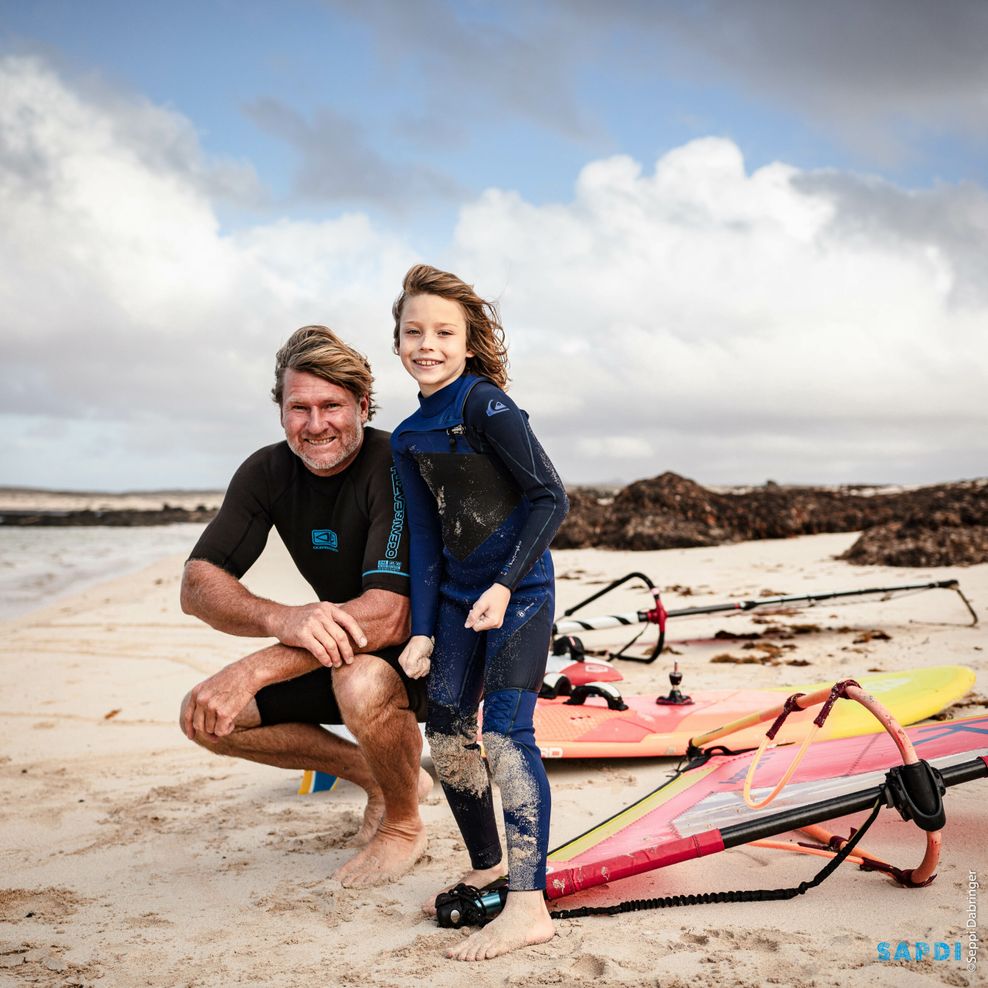 Björn Dunkerbeck und sein Sohn Liam am Strand, neben ihnen Surfbretter