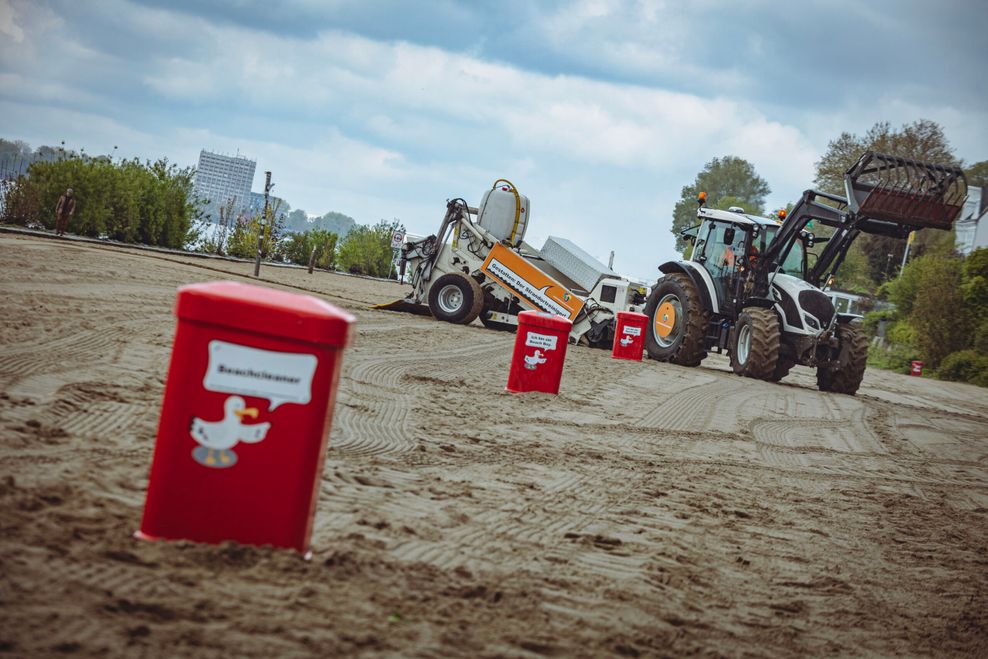 Elbstrand: rote Mülleimer und ein neuer „Beachcleaner“: ein Traktor zieht ein großes Reinigungsgerät.