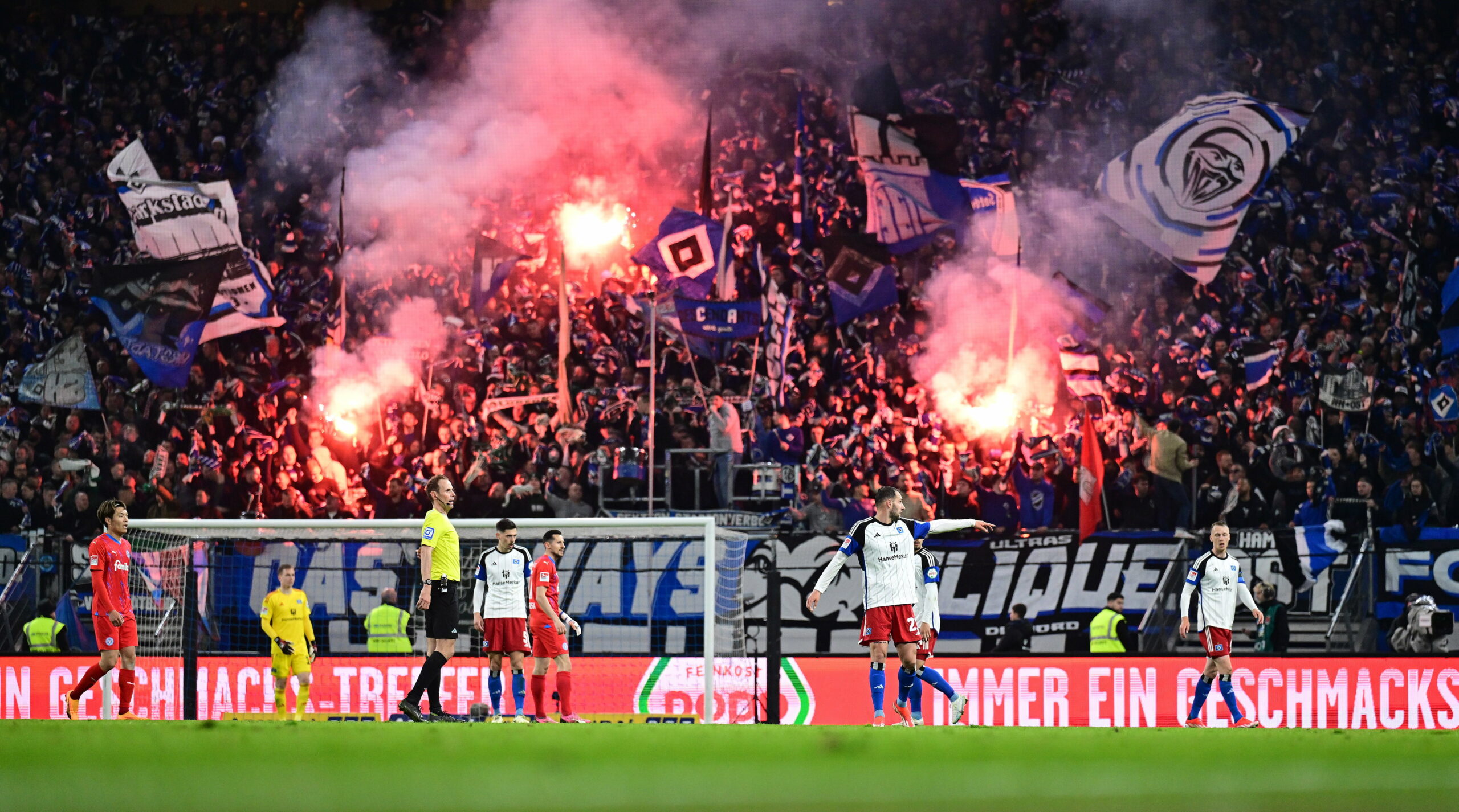 Auf der Nordtribüne im Volksparkstadion brennt Pyrotechnik, davor stehen HSV- und Kiel-Profis auf dem Platz