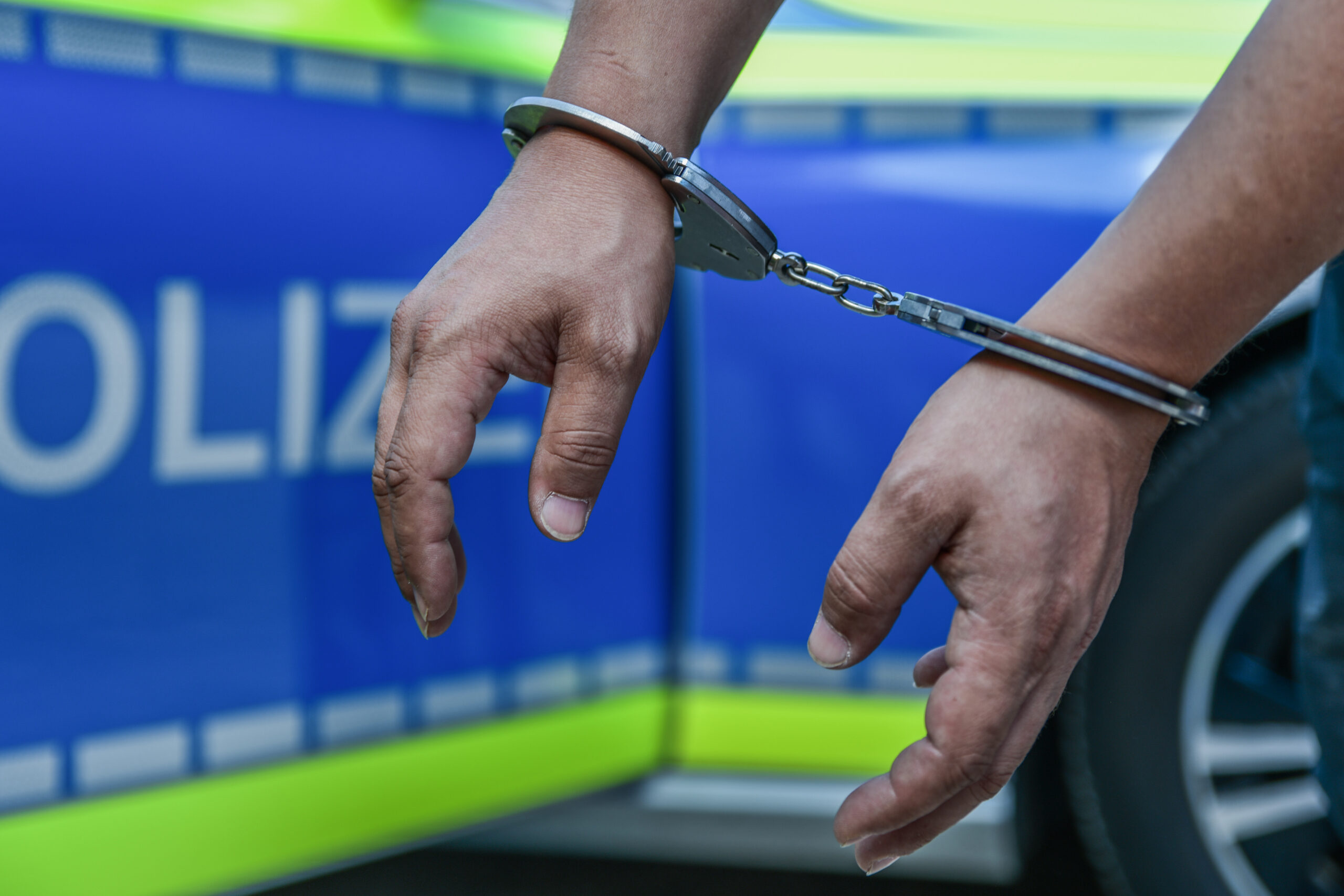 Hände in Handschellen vor einem Polizeiauto (Symbolbild)