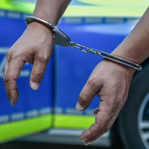 Hände in Handschellen vor einem Polizeiauto (Symbolbild)