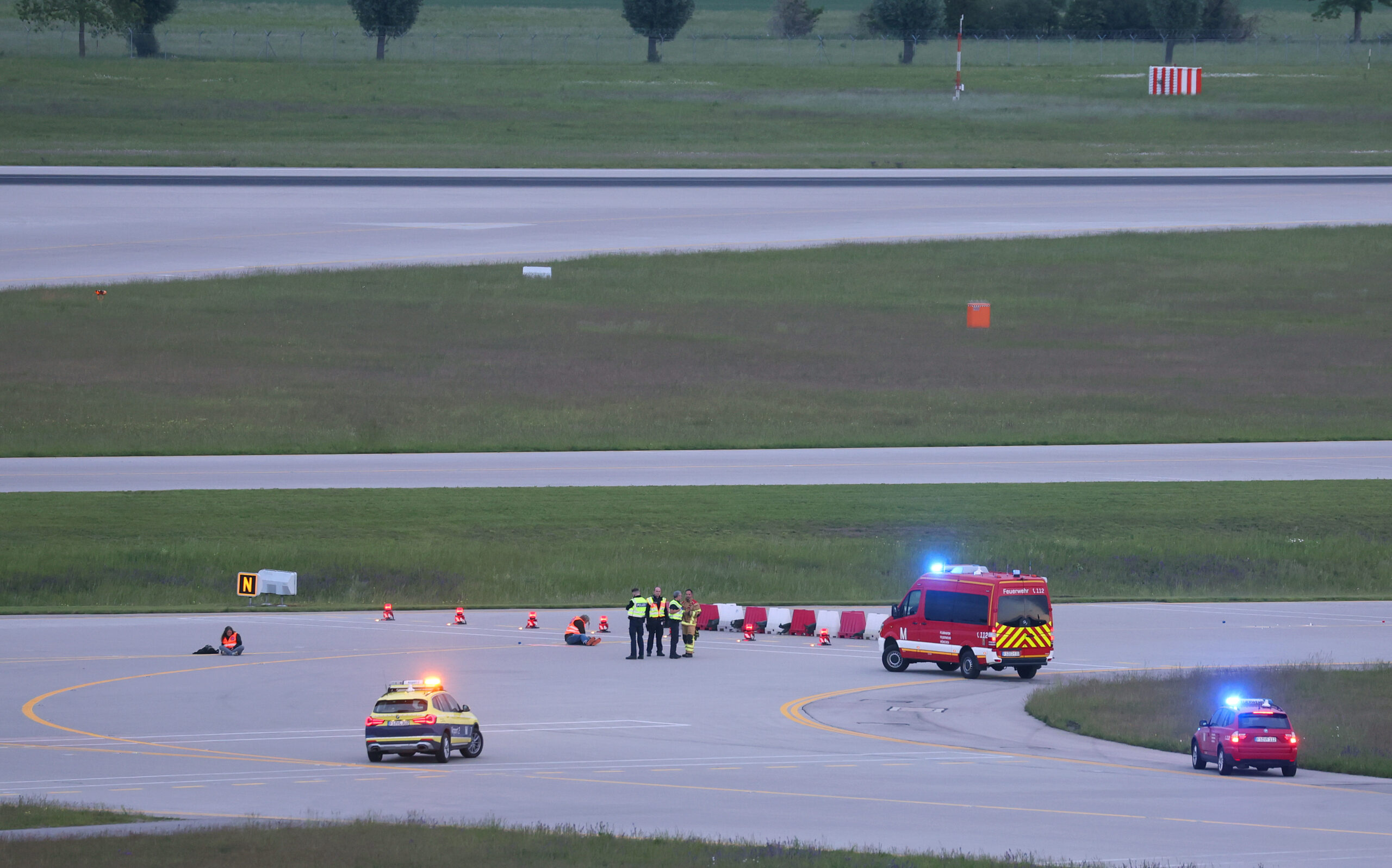 Einsatzkräfte von Polizei und Feuerwehr stehen auf einer Zufahrt für Start- und Landebahnen am Flughafen Franz-Josef-Strauß um Klimaaktivisten die sich dort festgeklebt haben.