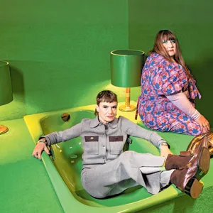 Zwei Frauen. Eine in einer grünen Badewanne sitzend die andere auf dem Rand sitzend. Vor grünem Hintergrund.