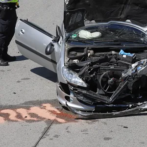 Ein Polizist steht neben dem Unfallwagen, die Front des Autos ist völlig zerstört, die Motorhaube steht offen.