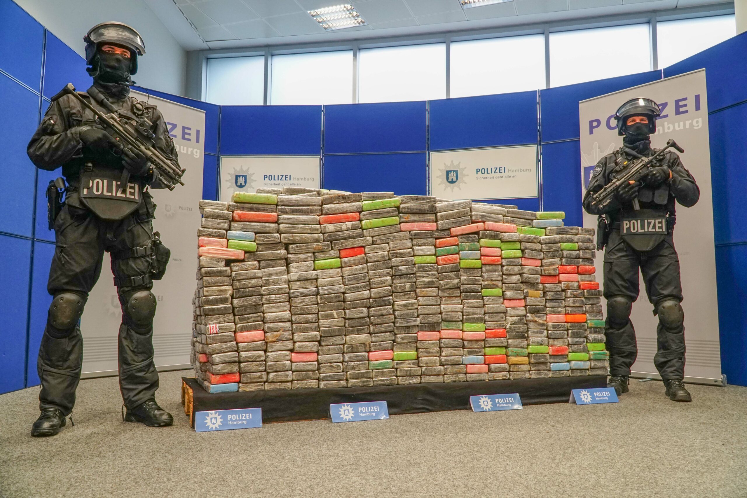 Zwei bewaffnete Polizisten stehen neben einer Palette, auf der sich sichergestellte Kokainpakete stapeln (Symbolfoto)