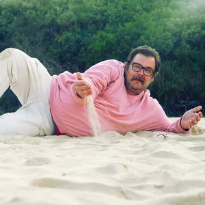 Erobique trägt eine helle Hose und einen rosafarbenen Pullover, liegt im Sand, den er durch eine Hand rieseln lässt