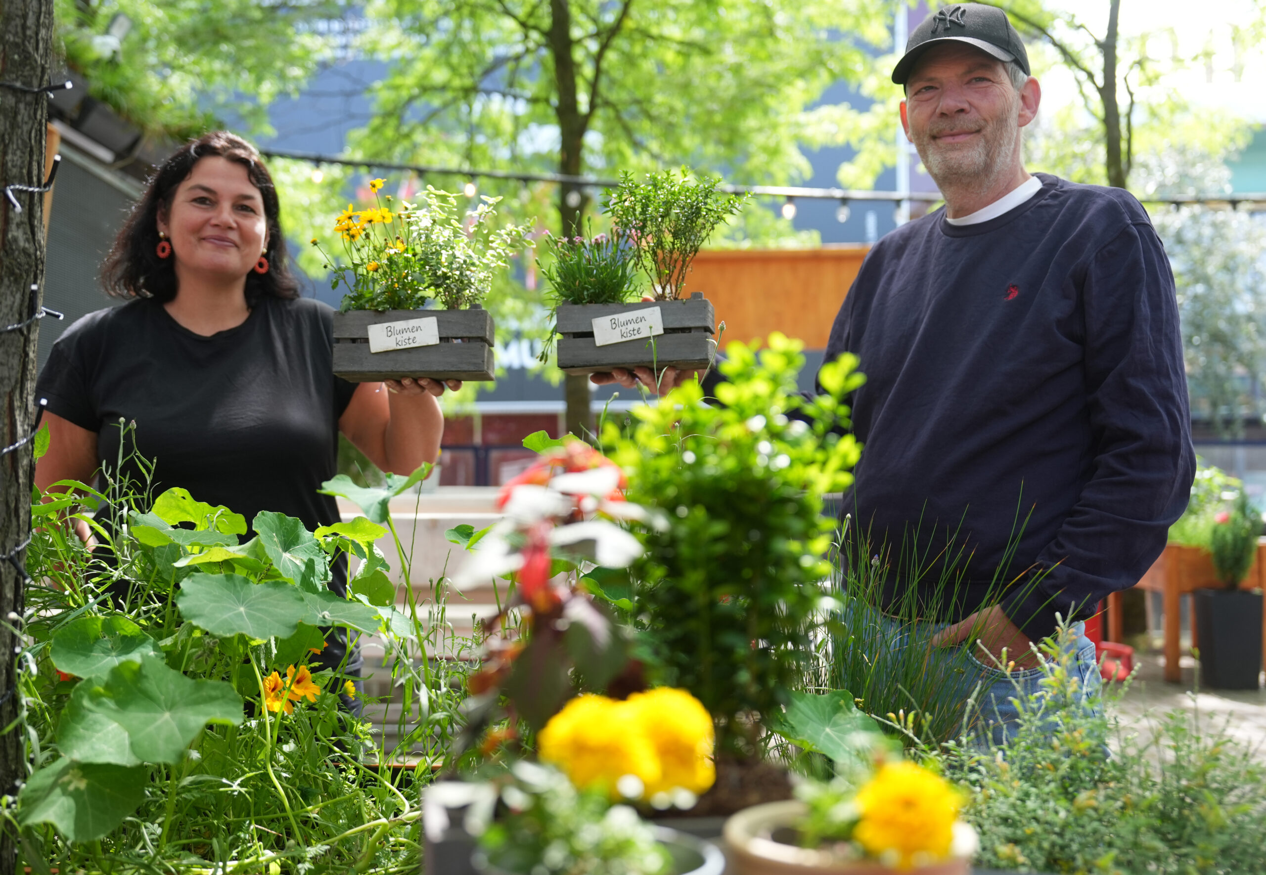 Sprecherin „Kiez Garden“, und Michael Sarnhof, Betreiber „Kiez Garden“, stehen mit Blumen hinter einem Hochbeet im Biergarten.