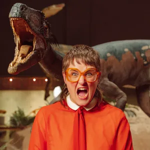 Eine junge Frau, kurze braune Haare, rote Brille, weite rote Bluse zieht eine Grimasse, hinter ihr eine Dinosaurier-Figur