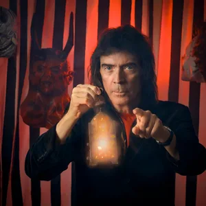 Er steht vor einer schwarzrot gestreiften Wand, hält eine Laterne in der einen Hand, zeigt mit der anderen in die Kamera. Im Hintergrund hängen drei Masken