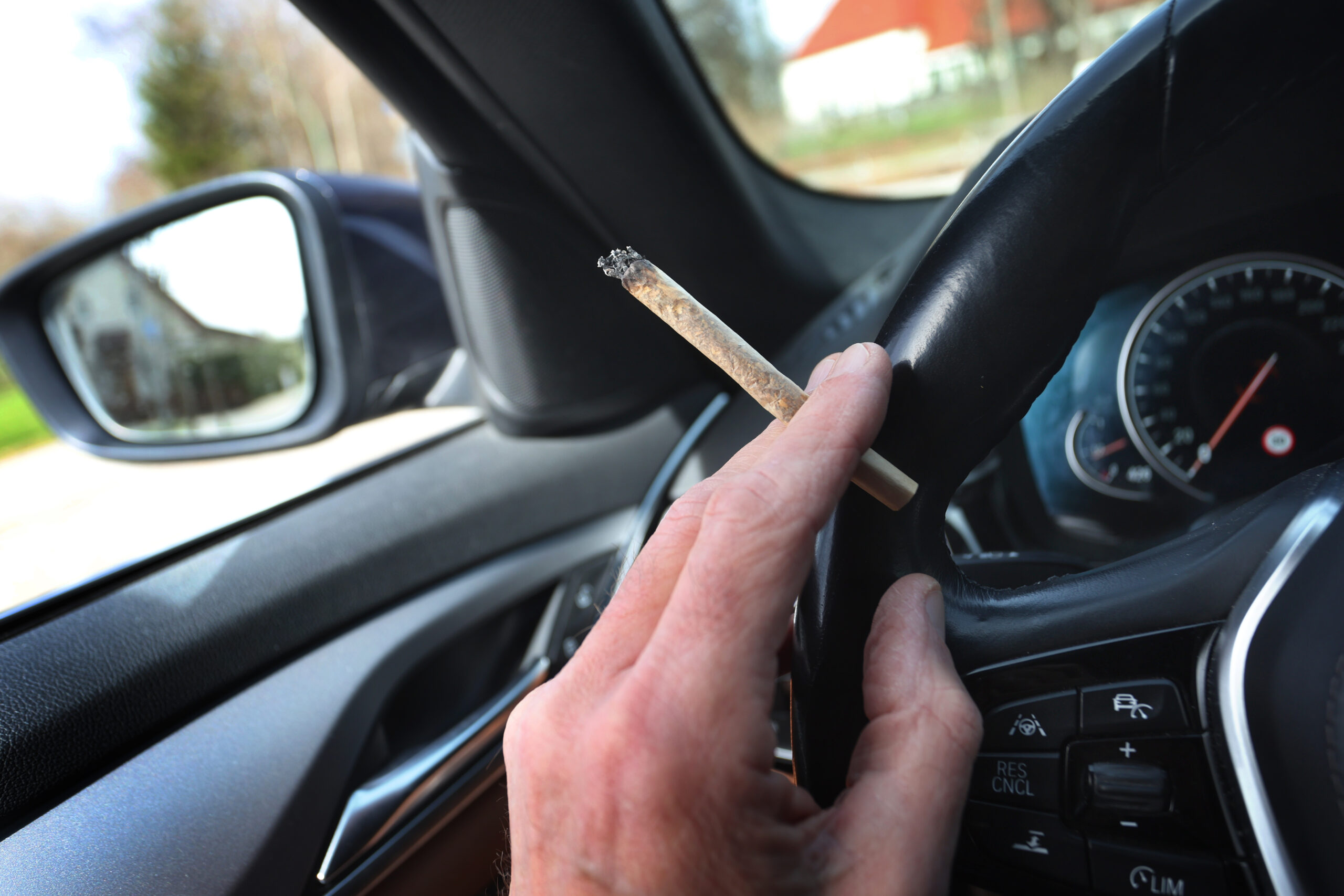 Ein Mann sitzt mit einem Joint zwischen den Fingern am Steuer eines Autos.