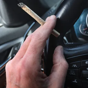 Ein Mann sitzt mit einem Joint zwischen den Fingern am Steuer eines Autos.
