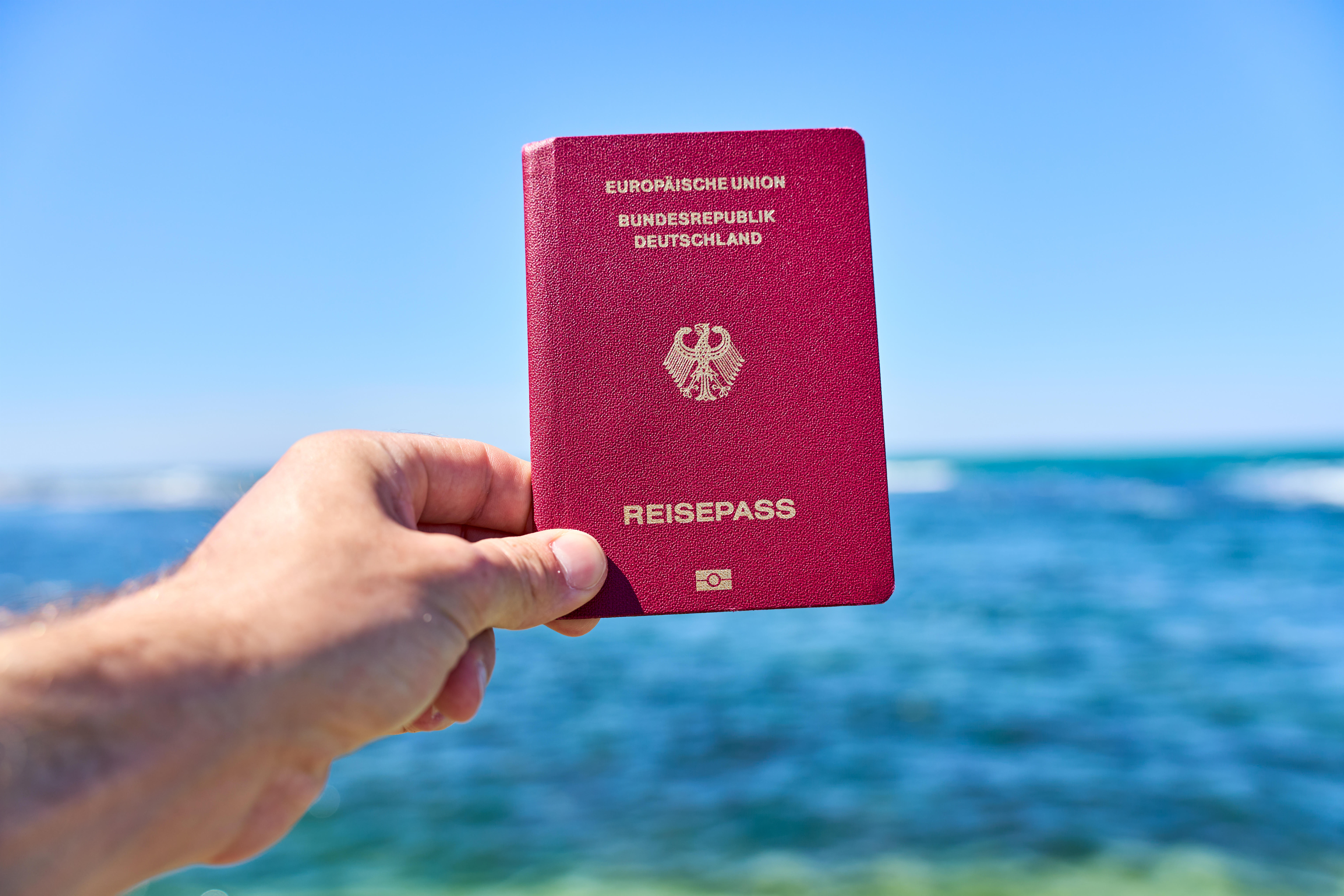 Ein Tourist hält einen deutschen Reisepass vor das Meer im Urlaub.