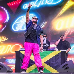 Bunter Bühnenhintergrund, vorne Sean Paul in einer pinken Hose und mit Sonnenbrille, hinter ihm ein DJ-Pult mit zwei Menschen