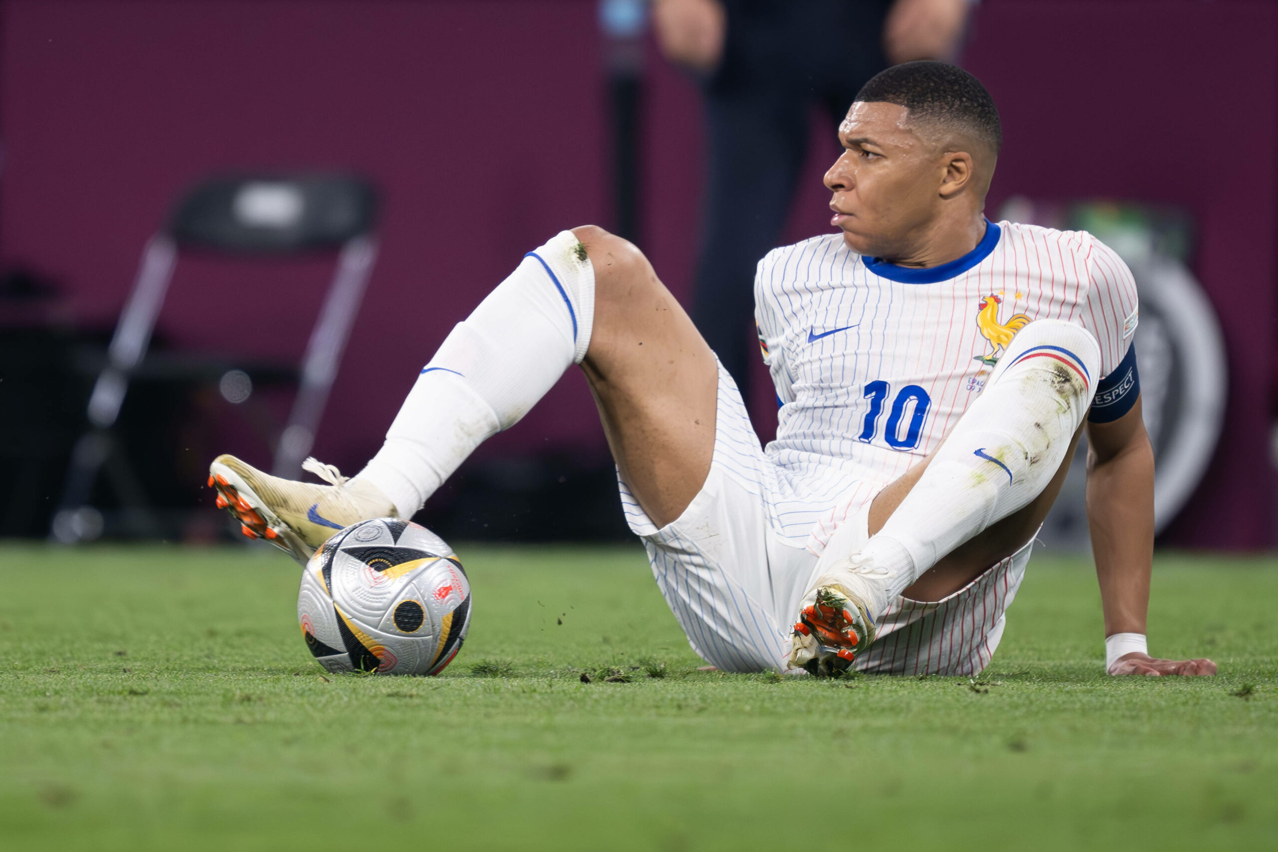 Kylian Mbappé sitzt auf dem Rasen, den Ball unter seinem Fuß eingeklemmt, sein Blick überrascht.