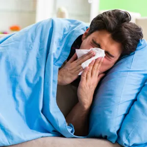 Ein Mann liegt mit Erkältung im Bett und putzt sich die Nase