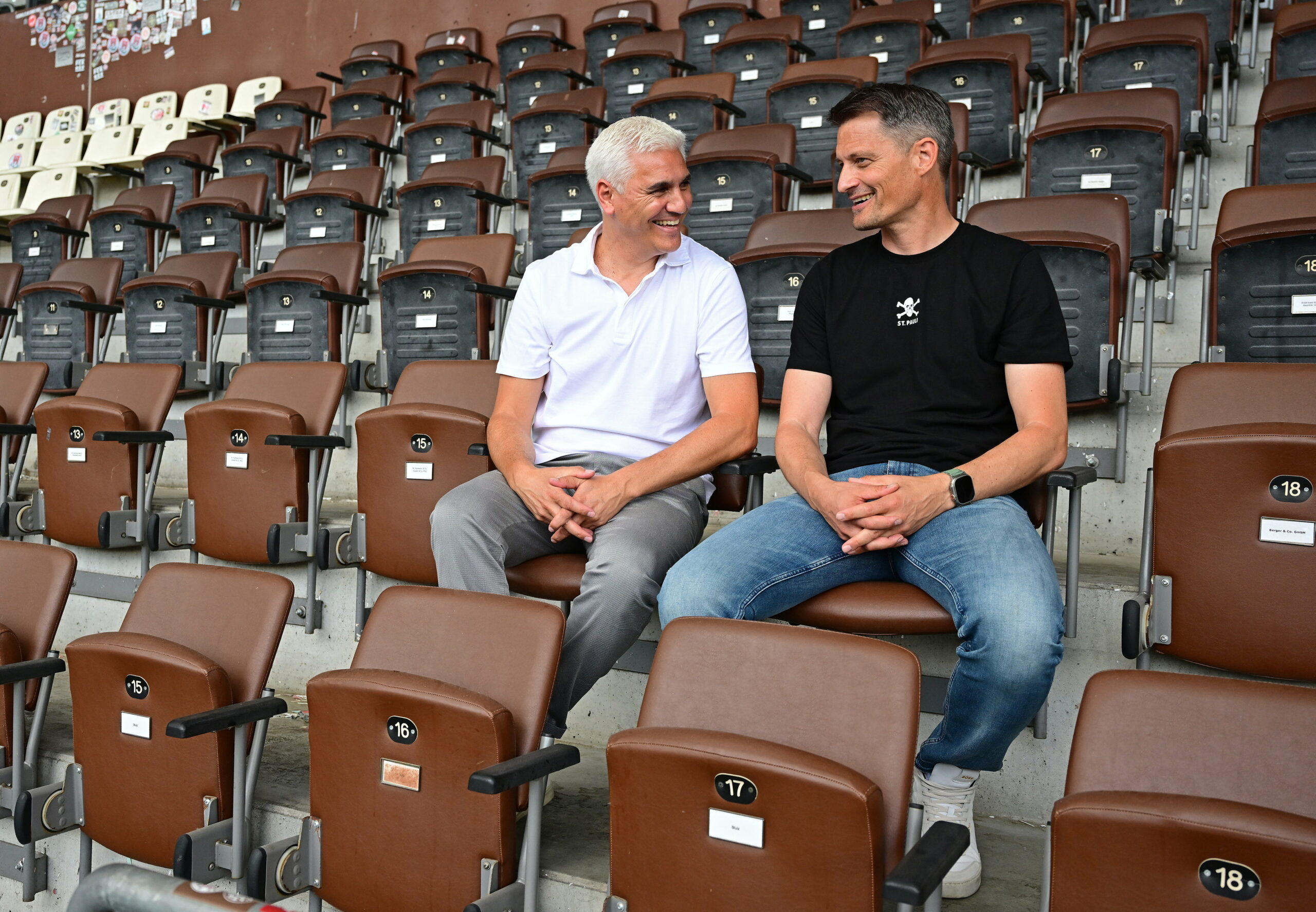 Sportchef Andreas Bornemann und Trainer Alexander Blessin sitzen auf der Tribüne und lachen