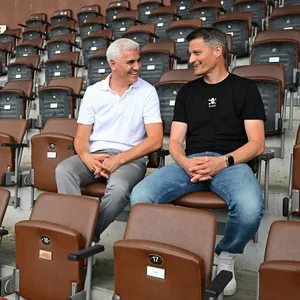 Sportchef Andreas Bornemann und Trainer Alexander Blessin sitzen auf der Tribüne und lachen