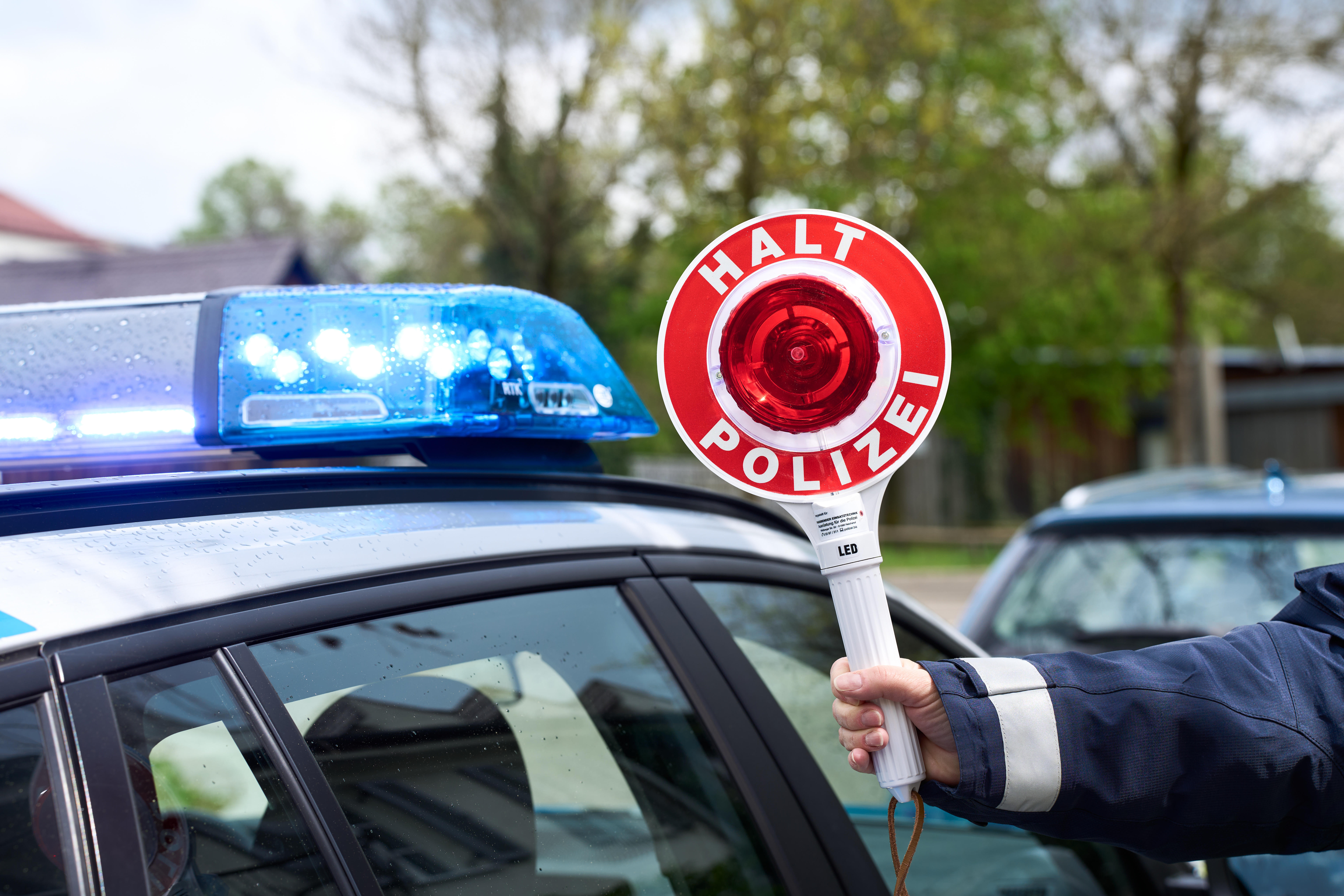 Polizist hält eine Polizeikelle mit Aufschrift Halt Polizei vor ein Polizeiauto mit Blaulicht