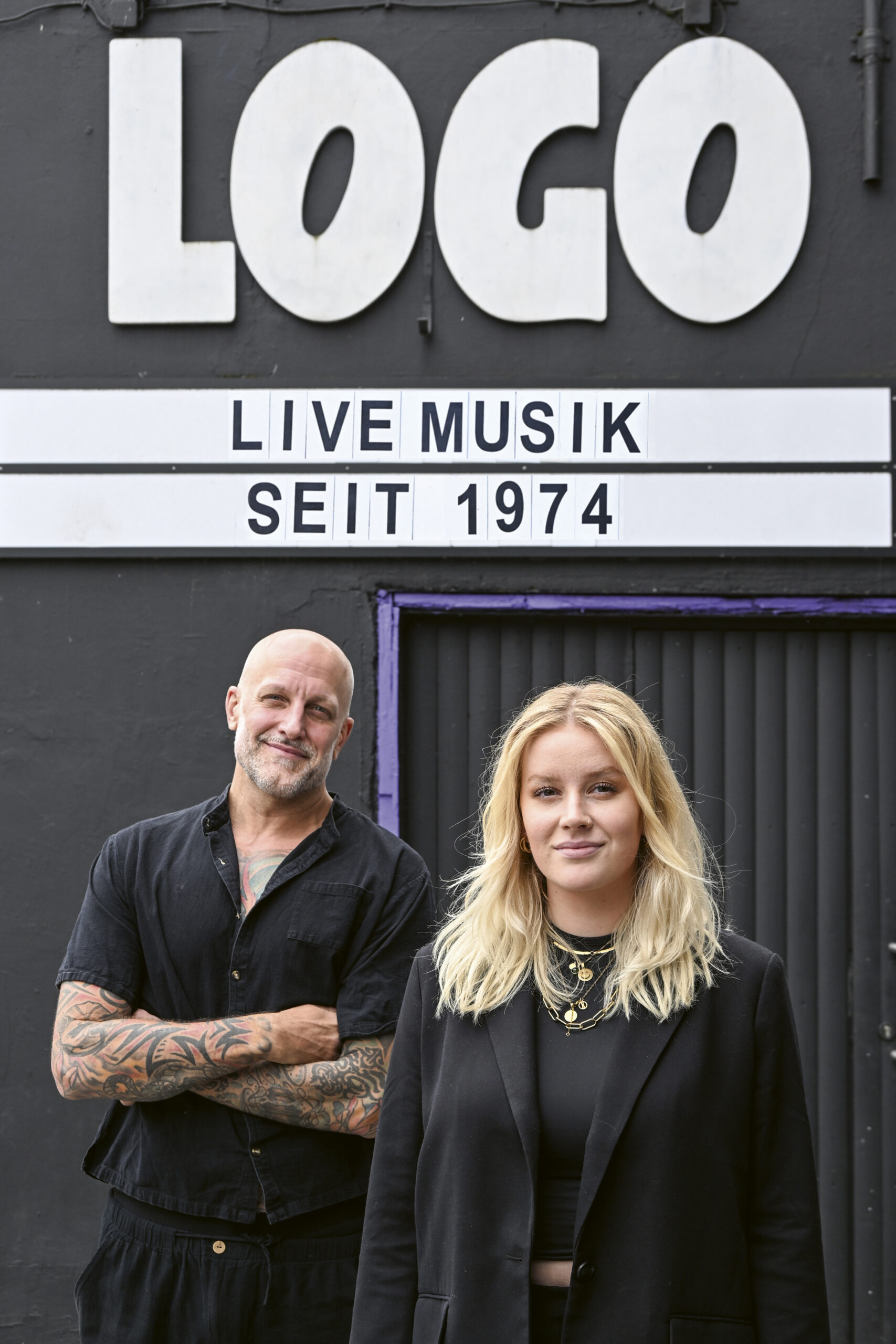 Chris August und Lea Goltz stehen in schwarzer Kleidung vor dem Logo