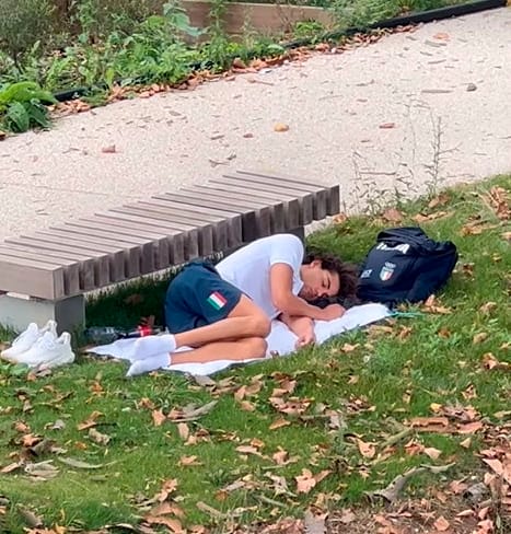 Thomas Ceccon schläft auf einem Handtuch vor einer Parkbank im Olympischen Dorf