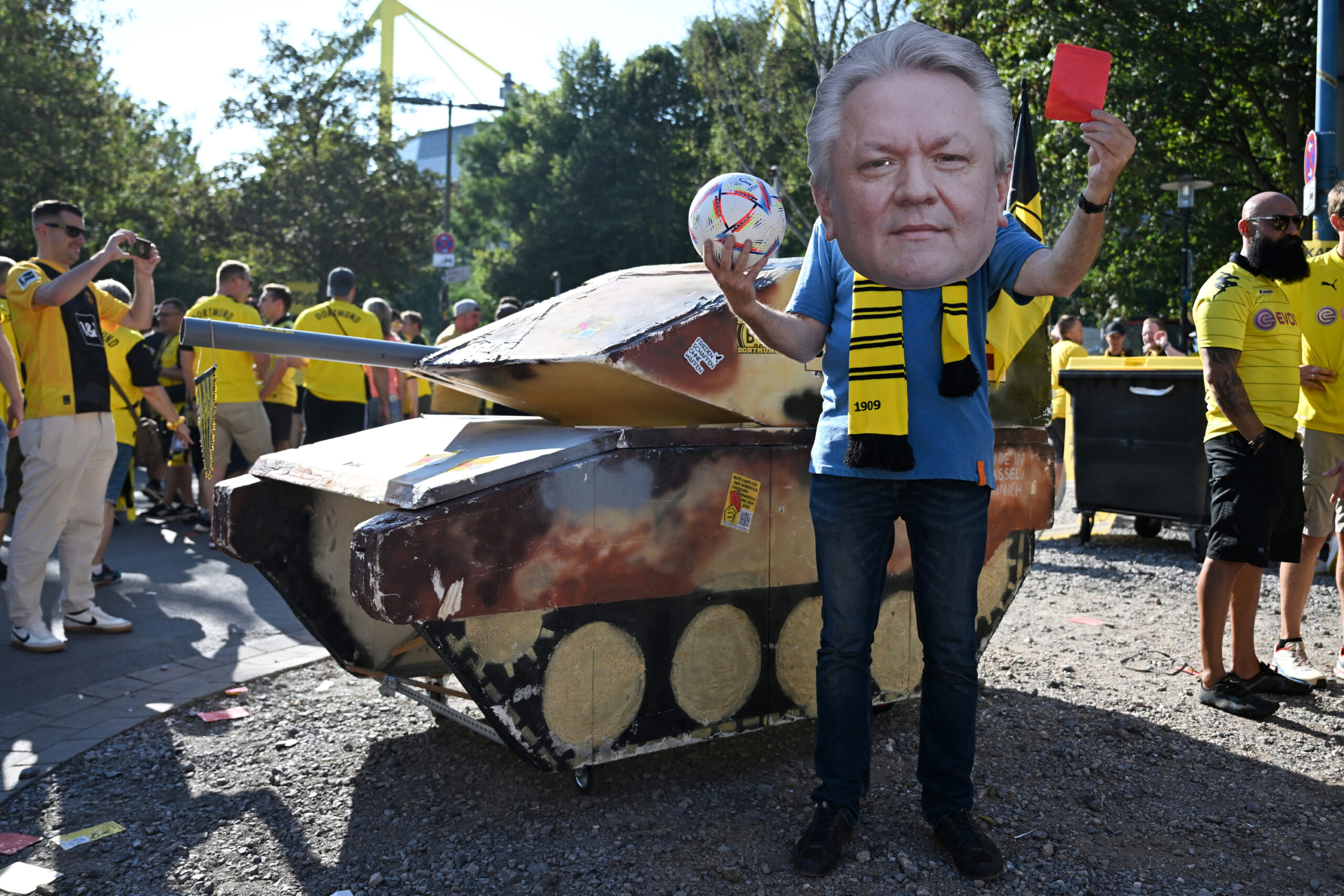 Eine Panzer-Attrappe steht neben einem Dortmund-Fan, der eine Maske mit dem Konterfei von Rheinmetall-Chef Armin Papperger trägt und dabei eine rote Karte hochhält.