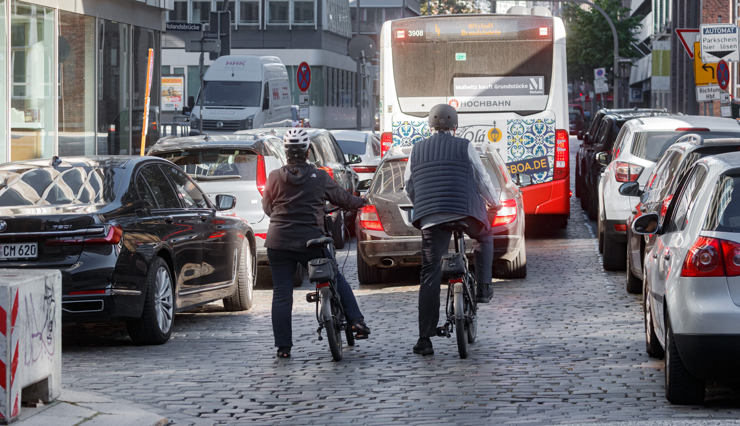 Ein Linienbus, ein Auto und zwei Radfahrer halten vor einer Ampel.