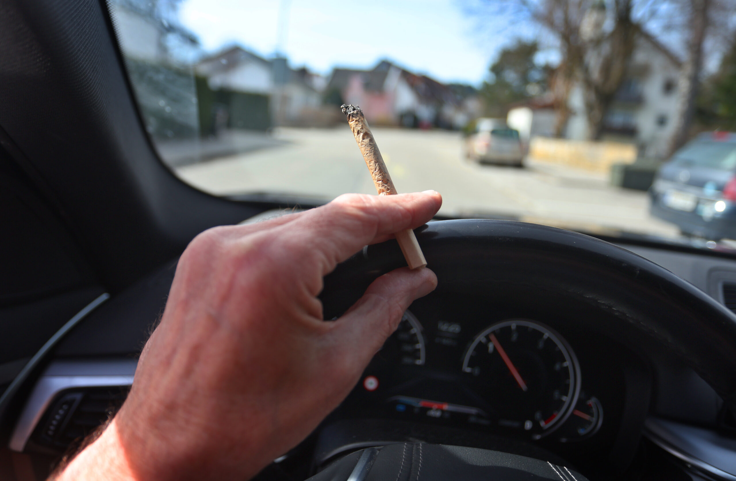 Ein Mann sitzt mit einem Joint zwischen den Fingern am Steuer eines Autos.