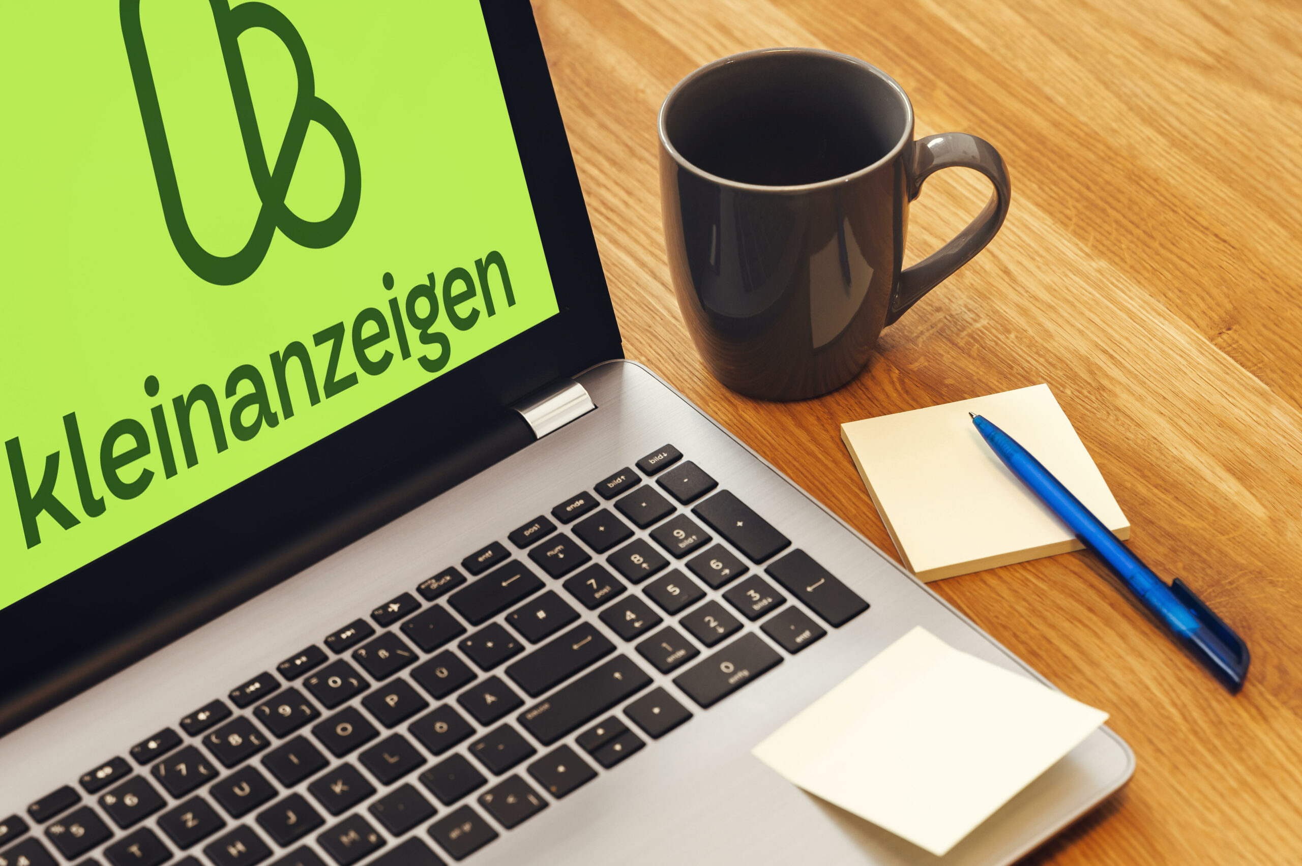 Laptop mit Kleinanzeigen-Logo steht neben Zettel, Stift und Tasse