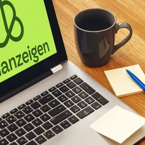 Laptop mit Kleinanzeigen-Logo steht neben Zettel, Stift und Tasse