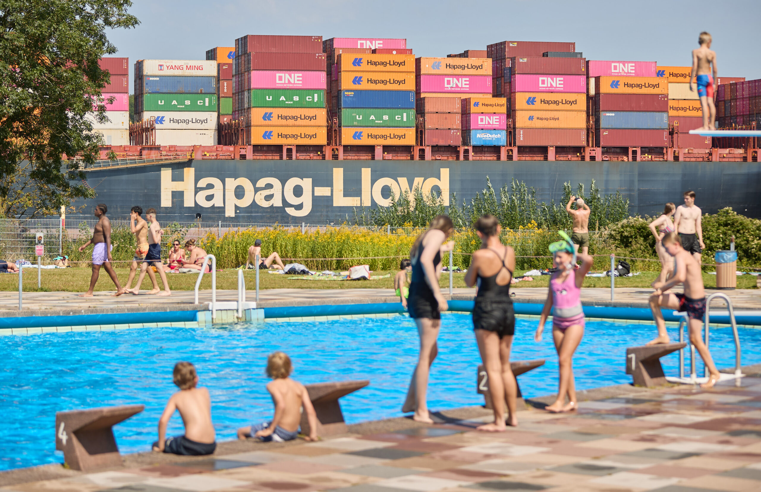 Das Containerschiff Colrado Express von Hapag-Lloyd fährt auf der Elbe flußaufwärts am Freibad vom Bäderland Finkenwerder vorbei.