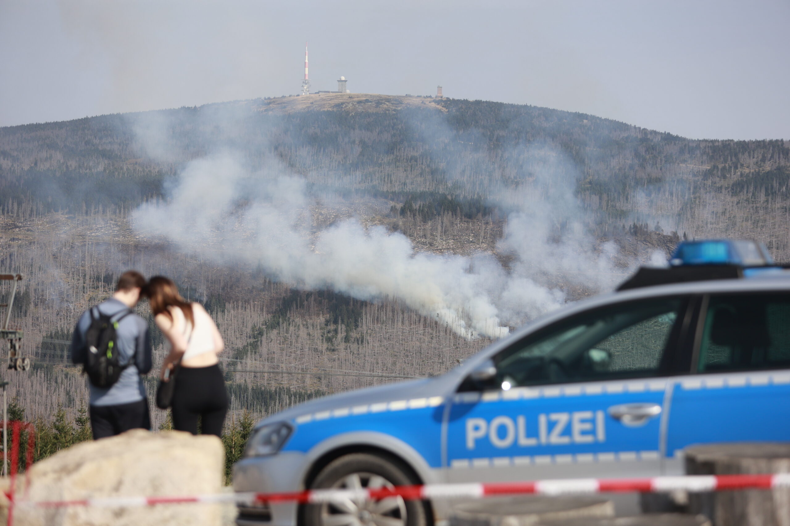 Rauchwolken steigen am Königsberg unterhalb des Brockens auf. Im Vordergrund steht ein Polizeifahrzeug an einer Absperrung.