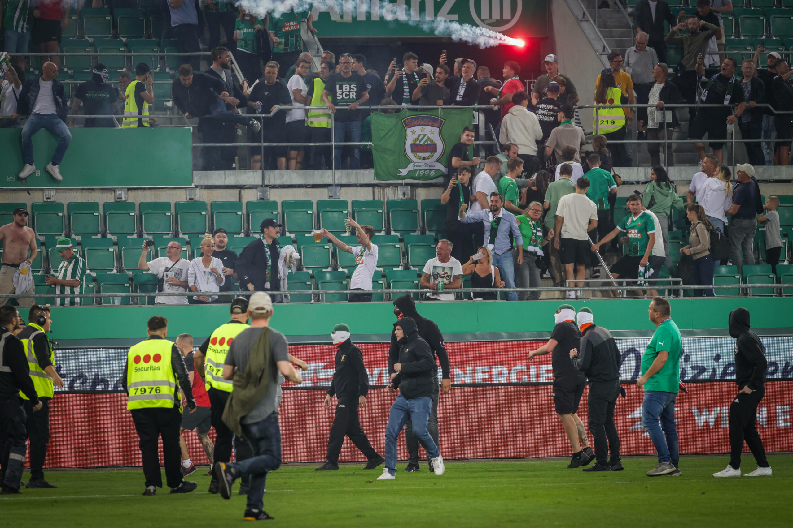 Teilweise vermummte Fans von Rapid Wien stehen auf dem Feld und werden von Ordner versucht aufzuhalten.Im Hintergrund fliegt eine Leuchtkugel in den Rapid Block