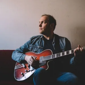 Er sitzt mit Gitarre auf einem Sofa und Blickt in Richtung eines Fensters