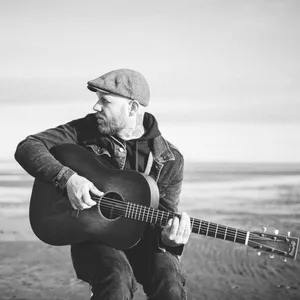 Matze Rossi am Meer, er sitzt auf einem Hocker, Gitarre in den Händen, und schaut zur Seite