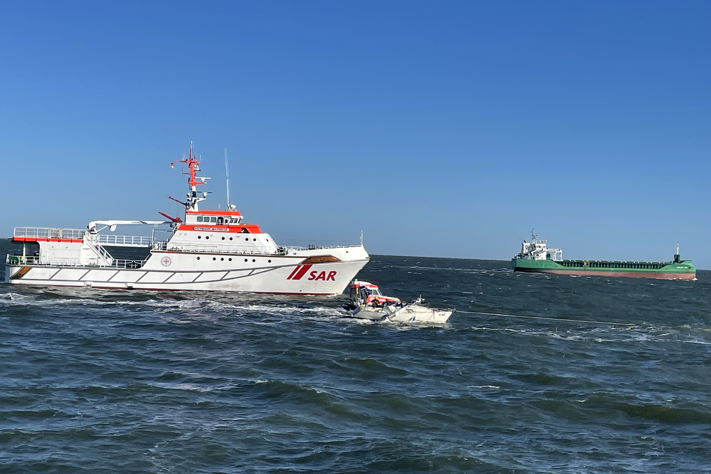 Eine kleine Segeljacht wird an einem Seil gezogen. Im Hintergrund sieht man ein Schiff der Seenotretter und einen Frachter.