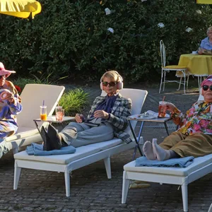 Schauspielerin Uschi Glas sitzt am Set ihrer neuen „Feel good“-Komödie auf einem Liegestuhl mit Sonnenbrille und Headset. Zu ihrer rechten und linken liegen ebenfalls zwei ältere Damen.
