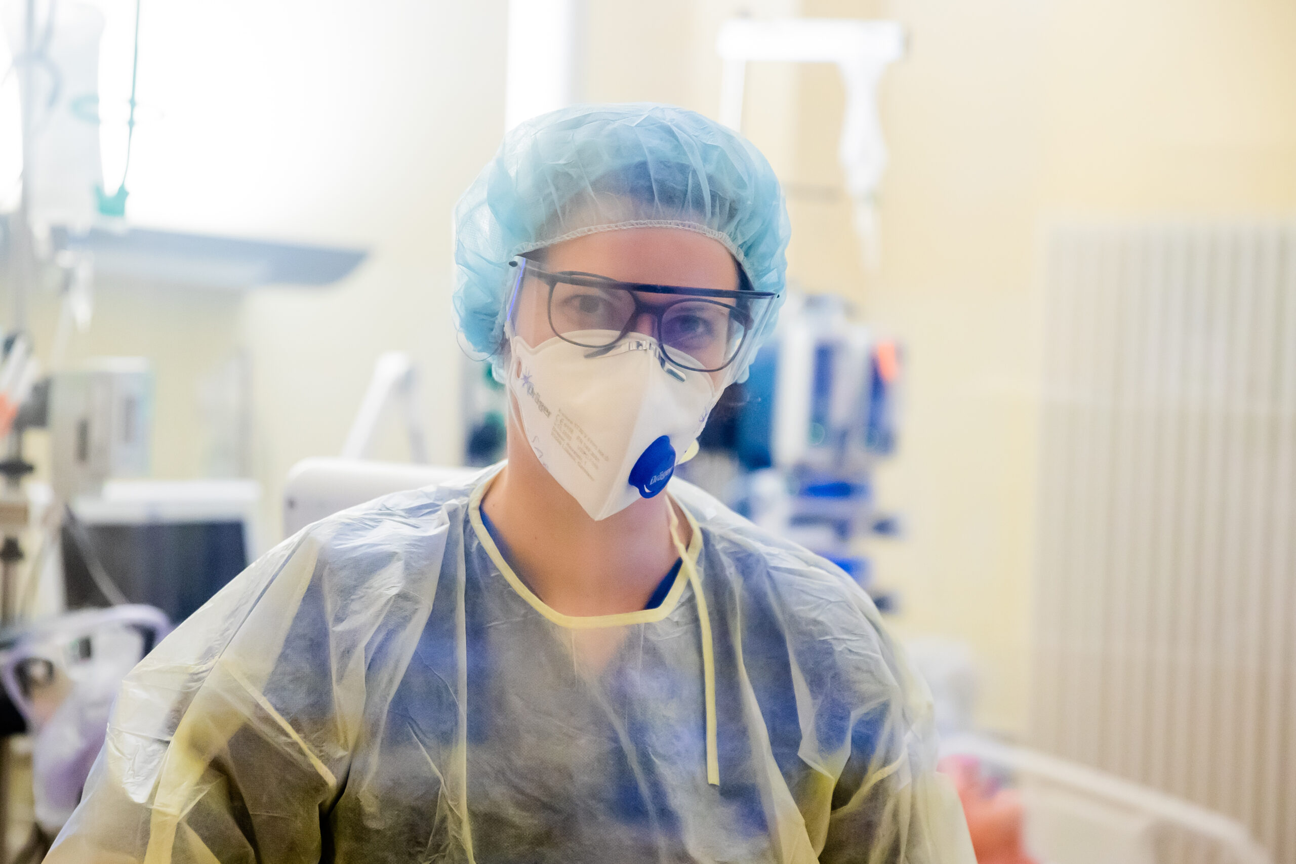 Eine Frau mit Kittel, Maske und Haube schaut in die Kamera. Im Hintergrund ist ein Krankenhauszimmer zu sehen.