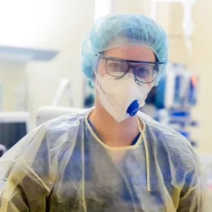 Eine Frau mit Kittel, Maske und Haube schaut in die Kamera. Im Hintergrund ist ein Krankenhauszimmer zu sehen.