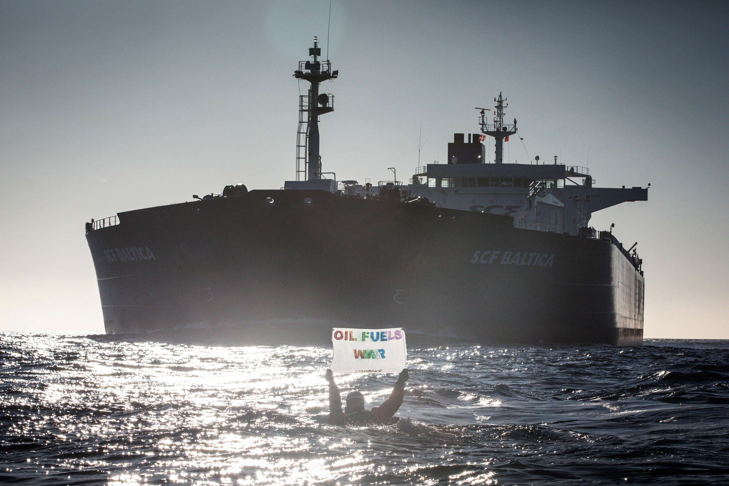 Eine Person – im Wasser schwimmend – hält ein Banner in die Höhe auf dem steht: „Oil Fuels War“. Im Hintergrund sieht man einen riesigen russischen Öltanker